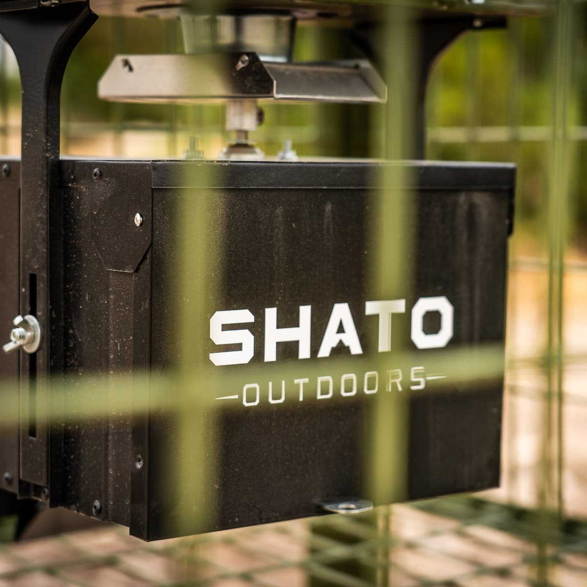 Close-up through the wire cage of the black control box under the hopper, marked SHATO OUTDOORS in white.
