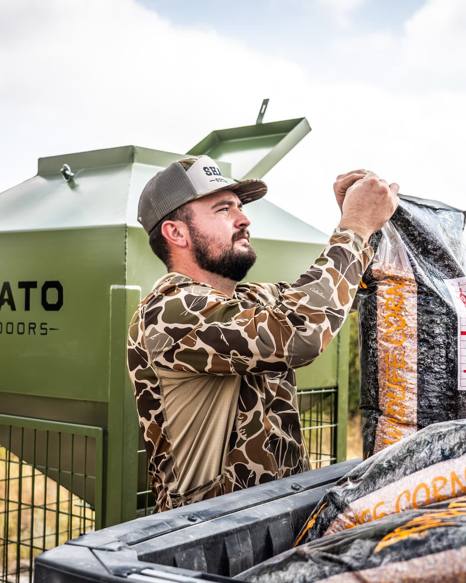 A man in a camo shirt and Shato cap lifts a bag of wildlife corn from a utility vehicle beside the feeder.